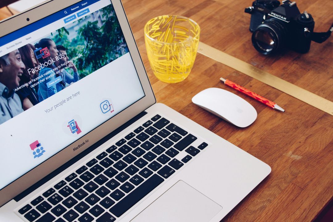 MacBook on a wooden desk showing the Facebook ads page, beside a camera, mouse and pen