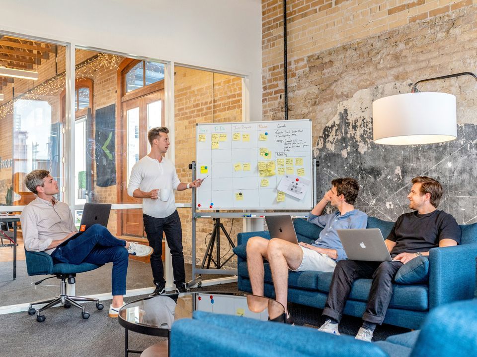 Four people in an office, one presenting at a whiteboard covered in sticky notes