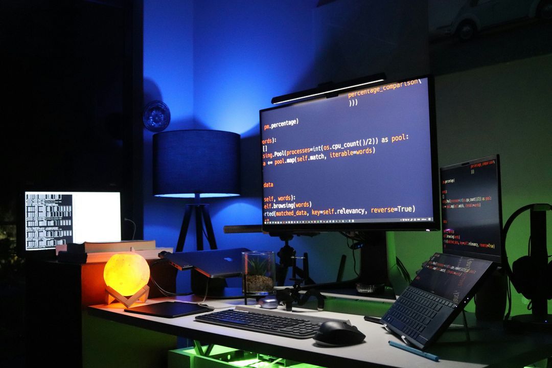 Desk setup lit in blue and green with a monitor and laptop showing Python scraping code
